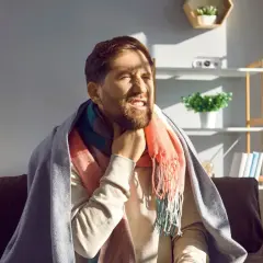 Mężczyzna z zarostem, okryty kocem i szalikiem, odczuwa ostry ból gardła przy przełykaniu, mimo braku gorączki.