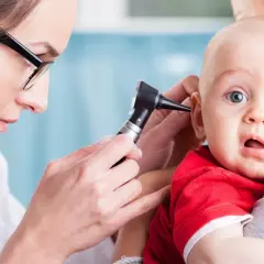 Lekarka bada ucho niemowlęcia, które może cierpieć z powodu bólu ucha. Dziecko ma szeroko otwarte oczy i usta.