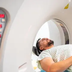 Mężczyzna leży w aparacie do tomografii komputerowej, przygotowując się do badania. Obok widoczny panel sterowania z cyfrowymi wskaźnikami.