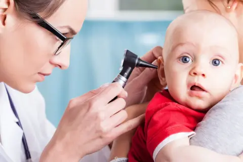 Lekarka bada ucho niemowlęcia, które może cierpieć z powodu bólu ucha. Dziecko ma szeroko otwarte oczy i usta.