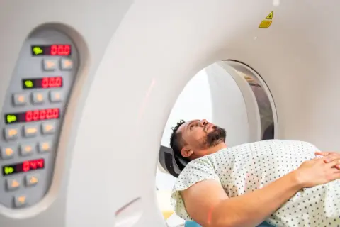 Mężczyzna leży w aparacie do tomografii komputerowej, przygotowując się do badania. Obok widoczny panel sterowania z cyfrowymi wskaźnikami.