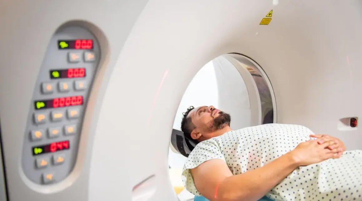 Mężczyzna leży w aparacie do tomografii komputerowej, przygotowując się do badania. Obok widoczny panel sterowania z cyfrowymi wskaźnikami.