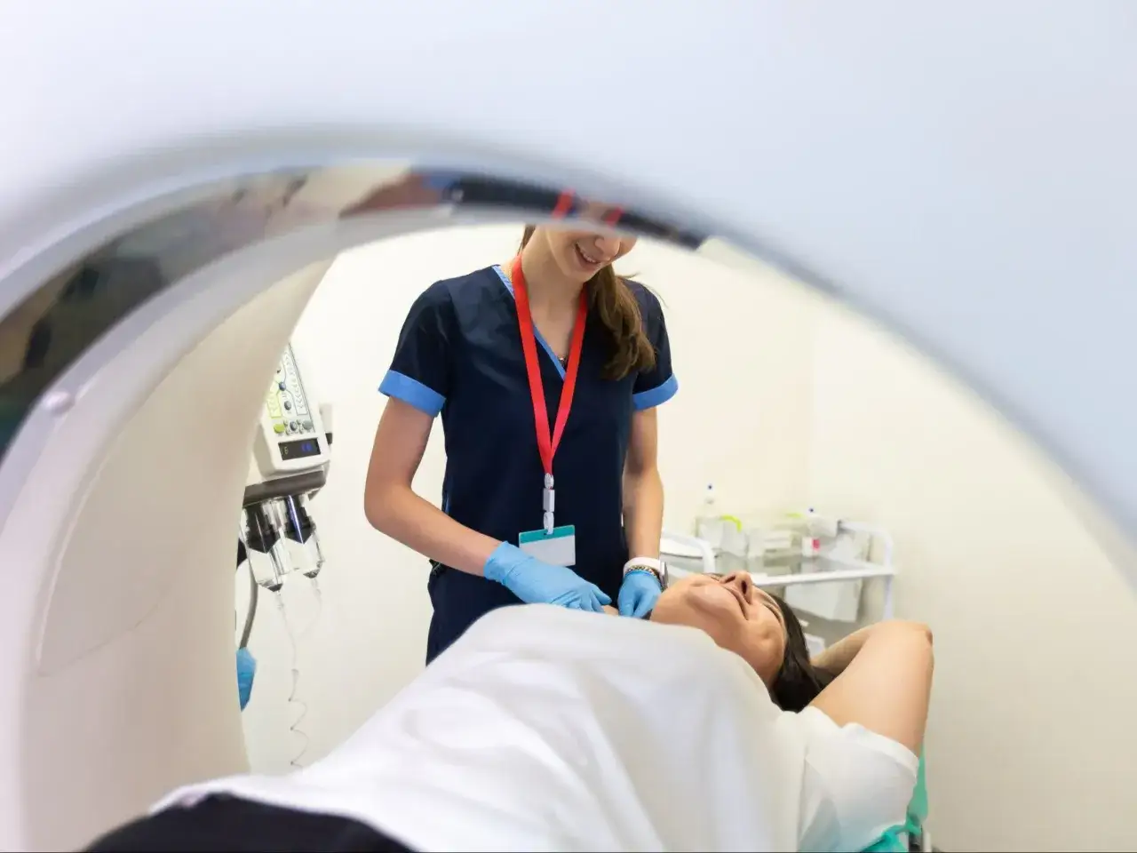Pacjentka leży na łóżku rezonansu magnetycznego, a pielęgniarka w niebieskich rękawiczkach przygotowuje ją do badania. To ważny objaw kliniczny.