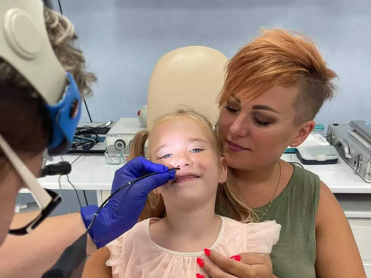 Mała pacjentka podczas endoskopii laryngologicznej, wspierana przez mamę. Lekarz w rękawiczce wprowadza endoskop.