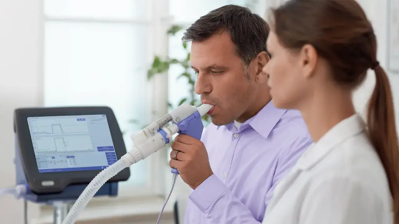 Mężczyzna wykonuje badanie spirometria, dmuchając w urządzenie pod nadzorem lekarki. Na ekranie widoczne są wykresy.