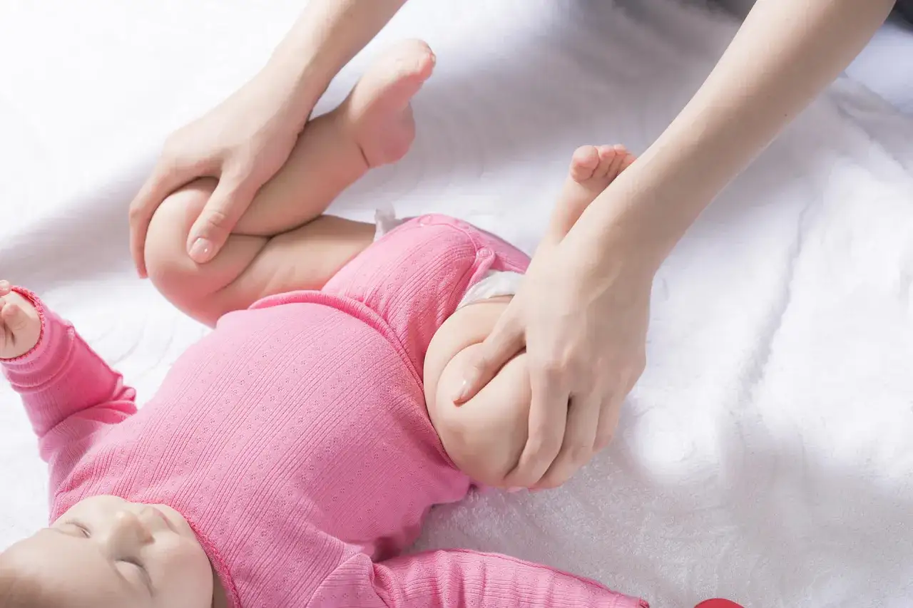 Delikatne ruchy dłoni pomagają w diagnostyce dysplazji stawu biodrowego u niemowlaka w różowym body.